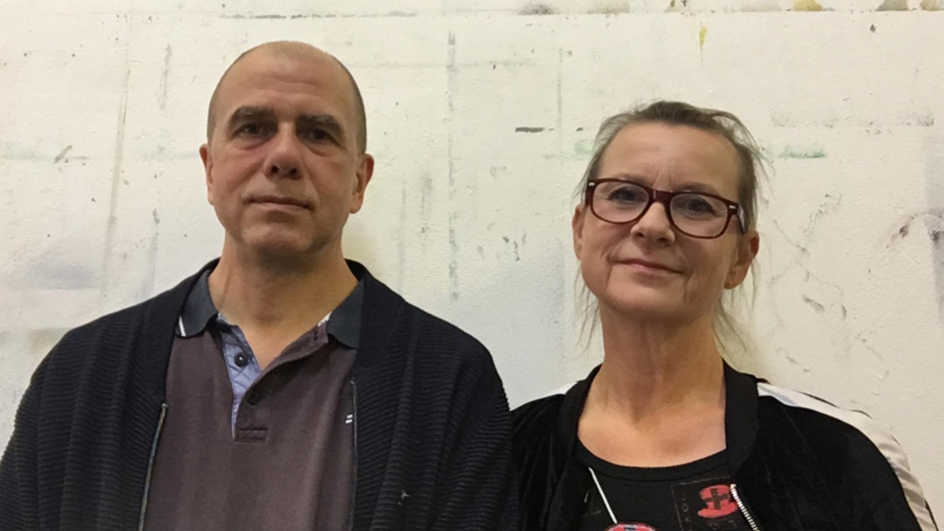 Ein Mann und eine Frau stehen vor einer grob verputzten Wand.