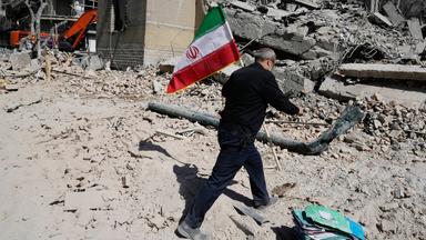 Ein Mann läuft durch den Schutt eines zerstörten Hauses und trägt die grün-weiß-rote Flagge Irans.