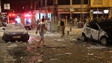 Eine Straßenkreuzung in Tel Aviv bei Nacht, nach einem Raketenangriff: Am Straßenrand stehen beschädigte Autos, Menschen begutachten die Schäden an Gebäuden und Straße.