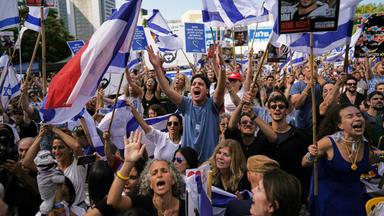 Auf einem Platz hat sich eine Menschenmenge versammelt, die weiß-blaue Fahnen schwenkt und jubelt.