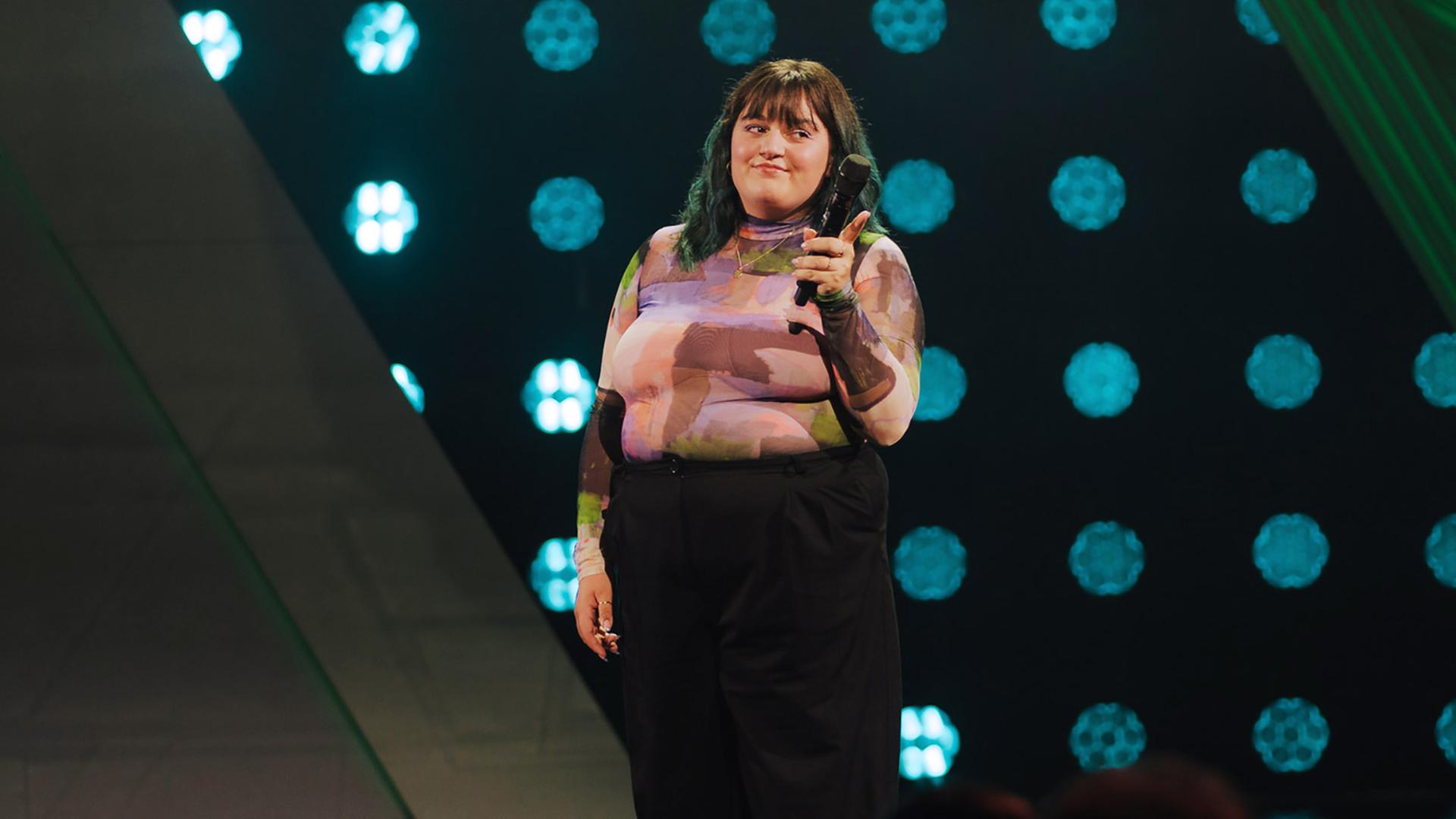 Teresa Reichl steht in der Mitte des Bildes. In der linken Hand hält sie ein Mikrofon und streckt einen Finger nach vorne.