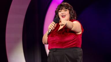 Teresa Reichl steht in der Mitte der Bühne. Sie streckt ihren linken Arm nach vorne aus. In der rechten Hand hält sie ein Mikrofon. Ihr Oberteil ist rot und sie trägt eine schwarze Hose.