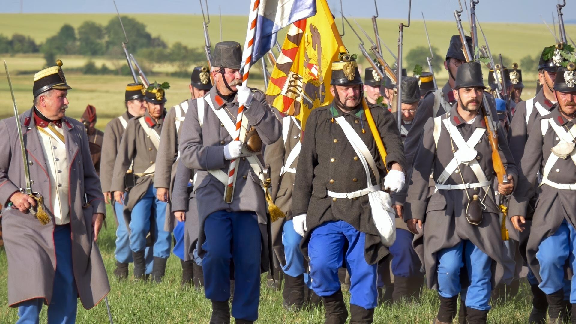 Das Bild zeigt eine Gruppe von Soldaten in historischen Uniformen, die in einer Reihe marchieren. Die Soldaten tragen graue Jacken mit weißen und schwarzen Akzenten sowie blaue Hosen. Einige von ihnen halten Gewehre, während ein Soldat eine Flagge mit sich führt. Die Szenerie ist grün und ländlich, mit sanften Hügeln im Hintergrund und einer klaren, blauen Himmel. Die Soldaten scheinen Teil einer Nachstellung der Schlacht von Königgrätz zu sein, die während des Österreichisch-Preußischen Krieges stattfand.