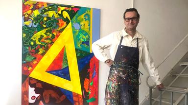 Ein Mann mit Brille und Schürze sützt sich an einem Treppengelände und steht neben einem Gemälde.