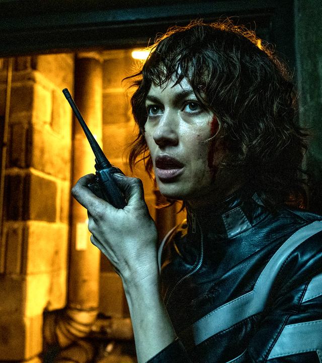 Der Kurier (Olga Kurylenko) steht in einem dunklen Keller und spricht in ein Walkie-Talkie.