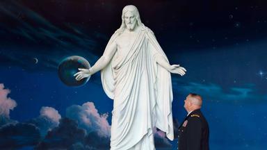 Ein US-Soldat in Uniform steht ehrfürchtig vor einer überlebensgroßen weißen Statue von Jesus Christus vor einem kosmischen Hintergrund mit Sternen und Planeten.