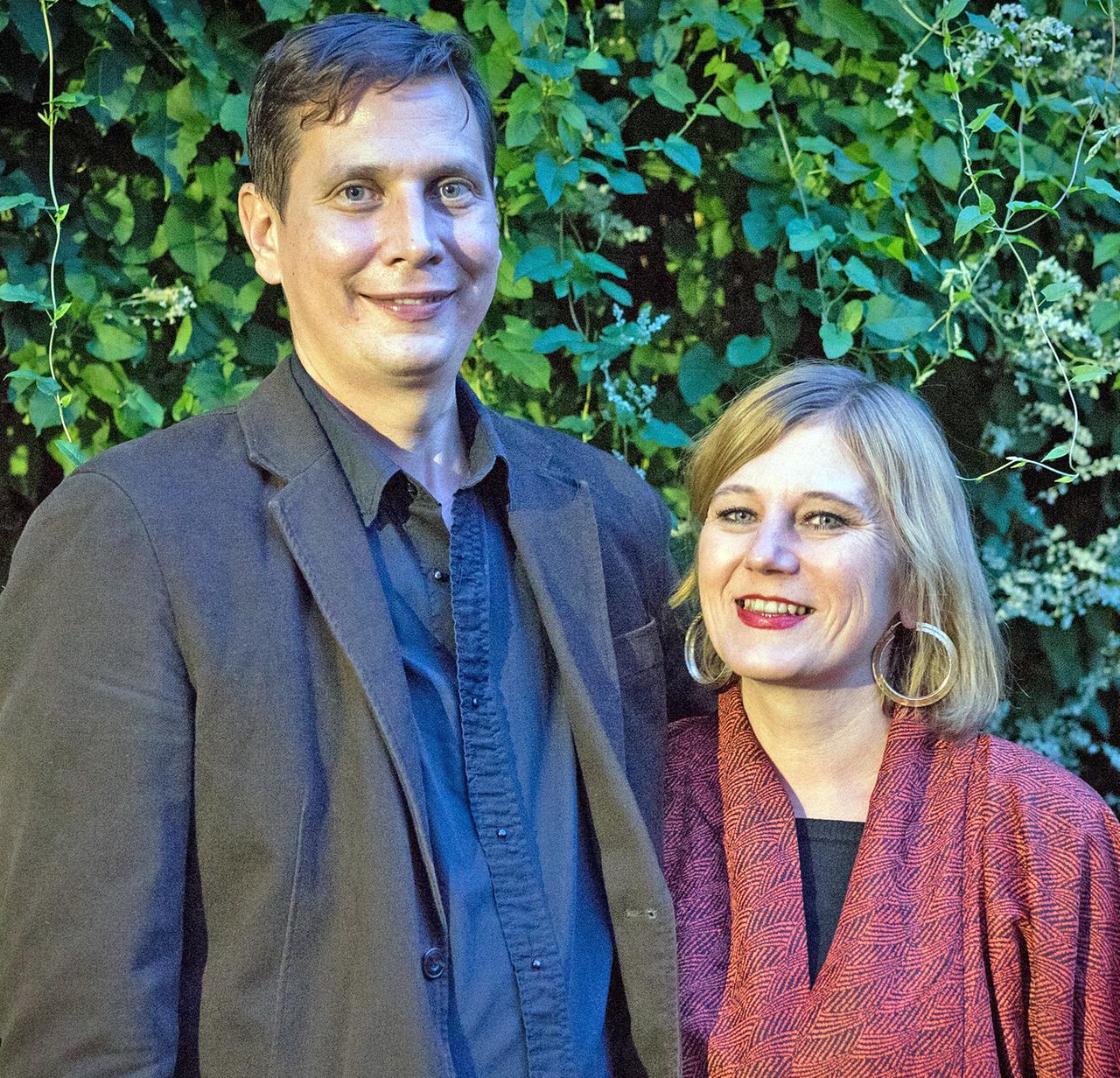 Michael Beamish und Jasmin Herold stehen eng nebeneinander vor einem Hintergrund aus grünen Rankpflanzen. Beide lächeln in die Kamera.