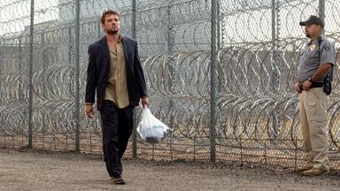 The Locksmith  Der Spezialist: Miller (Ryan Phillippe) läuft mit einer gefüllten Plastiktüte und ernstem Blick an einem hohen Maschendrahtzaun mit Stacheldraht entlang, während ein uniformierter Polizist in der Nähe steht.