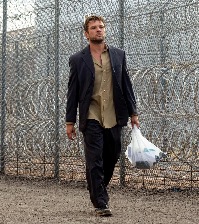 The Locksmith  Der Spezialist: Miller (Ryan Phillippe) läuft mit einer gefüllten Plastiktüte und ernstem Blick an einem hohen Maschendrahtzaun mit Stacheldraht entlang, während ein uniformierter Polizist in der Nähe steht.