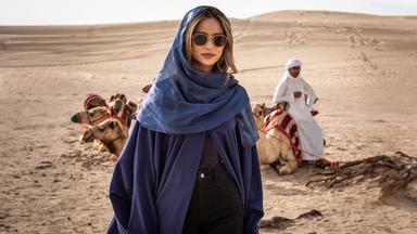 Eine Frau in dunklem Umhang und Sonnenbrille steht in einer Wüste vor einer Reihe sitzender Dromedare. Im Hintergrund sitzt ein Mann in weißer Kleidung auf einem Dromedar.