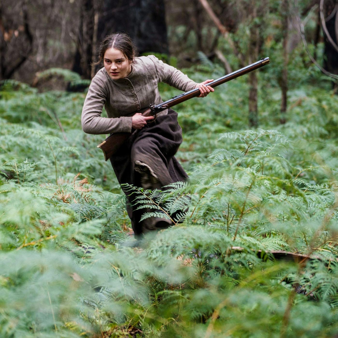 Clare (Aisling Franciosi) läuft entschlossen mit einem Gewehr durch eine bewaldete Lichtung voller Farne  Szene aus dem Film The Nightingale.