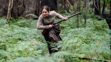 Clare (Aisling Franciosi) läuft entschlossen mit einem Gewehr durch eine bewaldete Lichtung voller Farne  Szene aus dem Film The Nightingale.