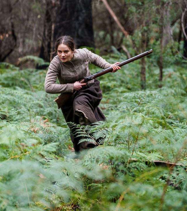 Clare (Aisling Franciosi) läuft entschlossen mit einem Gewehr durch eine bewaldete Lichtung voller Farne  Szene aus dem Film The Nightingale.