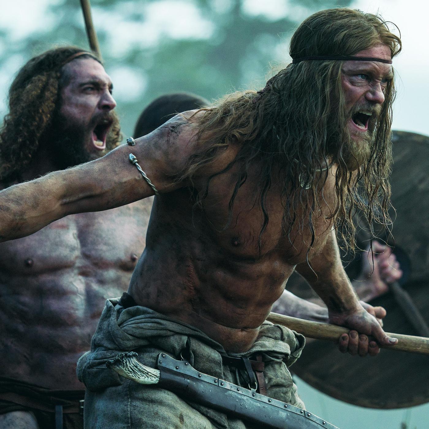Halbnahe: Amleth (Alexander Skarsgård), muskulös und oberkörperfrei, steht schreiend mit gezücktem Beil vor einem weiteren brüllenden Mann (Komparse) von ähnlicher Statur. Dahinter ist durch ein erhobenes Holzschild und eine Lanze eine weitere Person zu erahnen. Im Hintergrund sind unscharf Bäume zu erkennen.
