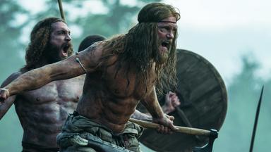 Halbnahe: Amleth (Alexander Skarsgård), muskulös und oberkörperfrei, steht schreiend mit gezücktem Beil vor einem weiteren brüllenden Mann (Komparse) von ähnlicher Statur. Dahinter ist durch ein erhobenes Holzschild und eine Lanze eine weitere Person zu erahnen. Im Hintergrund sind unscharf Bäume zu erkennen.