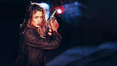 Eine Frau mit nassen Haaren und einem Lederjacke hält eine Pistole und schaut entschlossen. Der Hintergrund ist dunkel.
