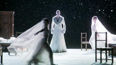 Auf einer Bühne mit weißem Boden und schwarzem weiß gesprenkelten Hintergrund laufen im Vordergrund zwei mit großen durchsichtigen Schleiern verhangene Frauen aufeinander zu, während in der Mitte eine Person in langem weißen Mantel steht.