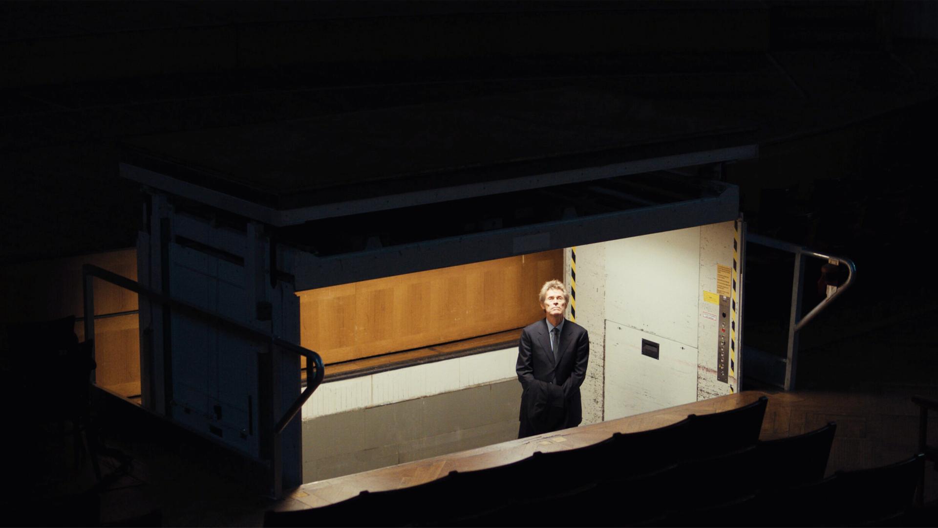 Blick von schräg oben in einen hell erleuchteten Bühnenaufzug. Darin steht ein Mann im Anzug und schaut nach oben.