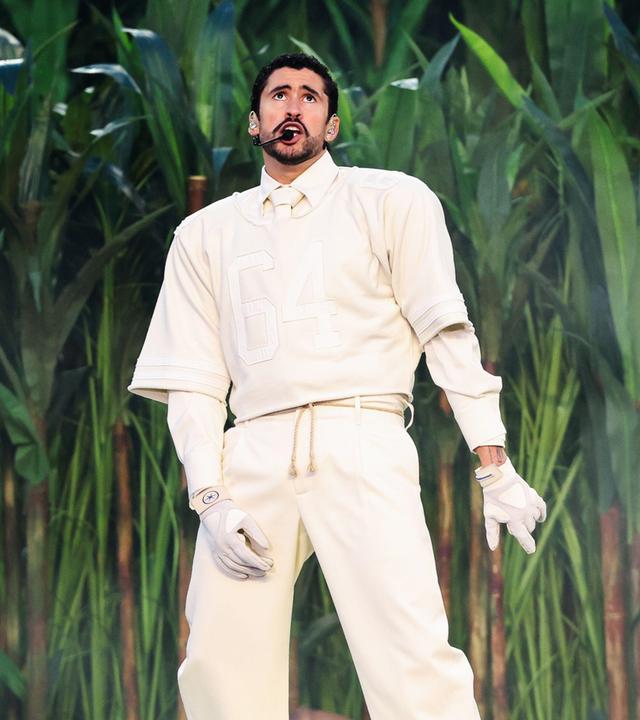 Bad Bunny performt vor einem Bühnenbild aus Zuckerrohrpflanzen. Er trägt weisse Kleidung und weiße Handschuhe.