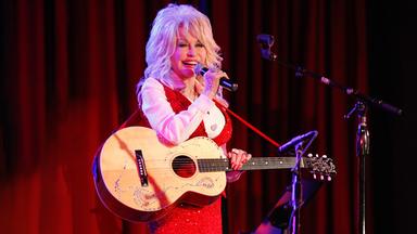 Dolly Parton hält in der linken Hand eine Gitarre. Sie steht vor einem roten Vorhang und singt in ein Mikrofon.