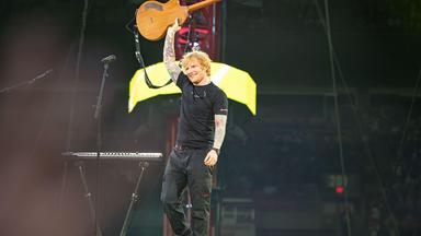 Ed Sheeran steht auf einer Konzertbühne und hält seine Akustikgitarre mit ausgestrecktem Arm über den Kopf. Der Sänger trägt ein schwarzes T-Shirt und eine schwarze Hose.