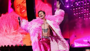 Harry Styles in pinkfarbener Metallic-Hose und Plüschmantel auf einer Bühne. Im Hintergrund Scheinwerfer und Übertragungsleinwand.