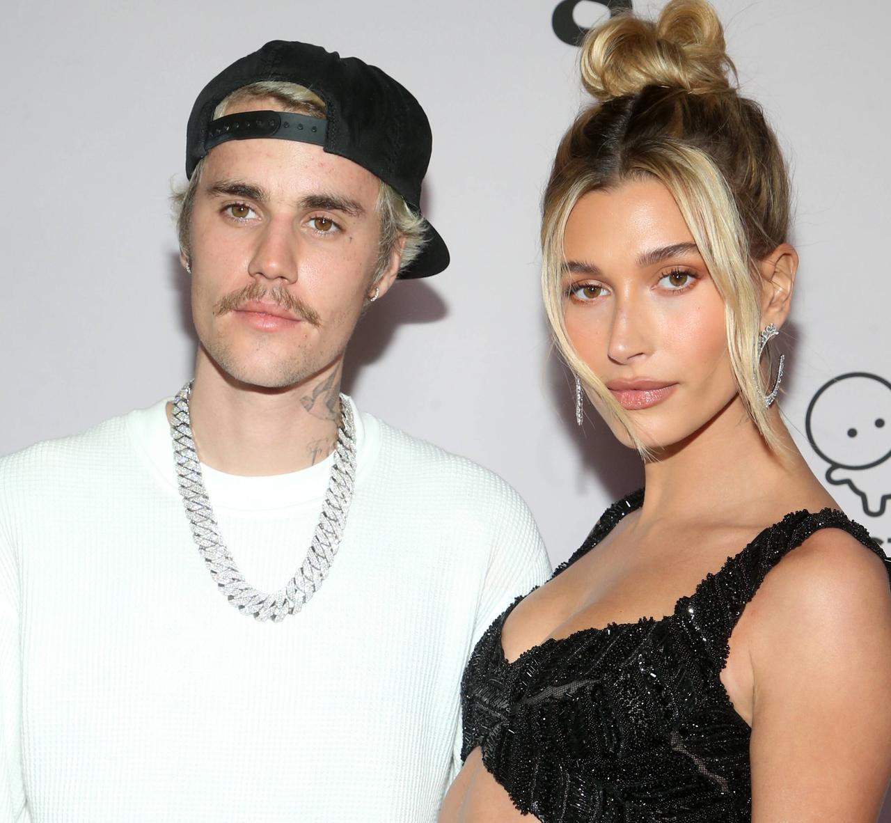Justin Bieber und Ehefrau Haily Bieber stehen vor einer hellen Wand, er trägt einen weißen Pullover und schwarze umgedrehte Basecap, sie ein schwarzes Bauchfreikleid.