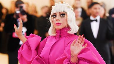 Lady Gaga trägt ein rosafarbenes Kleid mit Puffärmeln. Hellblonde Haare reichen ihr bis zur Schulter, und sie trägt lange künstliche Wimpern. Im Hintergrund sind Fotografen und ein Mann im Smoking zu sehen.