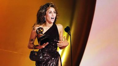 Miley Cyrus im Abendkleid auf Bühne vor Mikrofon, hält Auszeichnung in rechter Hand.