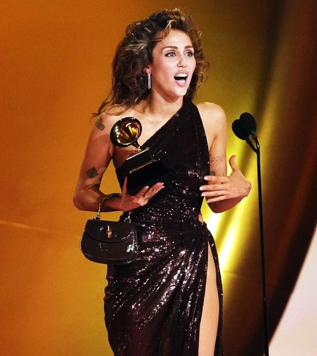 Miley Cyrus im Abendkleid auf Bühne vor Mikrofon, hält Auszeichnung in rechter Hand.