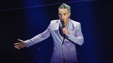 Robbie WIlliams steht auf einer Bühne, hält ein Mikrofon in der rechten Hand und streckt den linken Arm mit geöffneter Hand nach vorne aus. Er trägt einen hellgrauen Anzug und singt vor dunkelblauem Hintegrund.