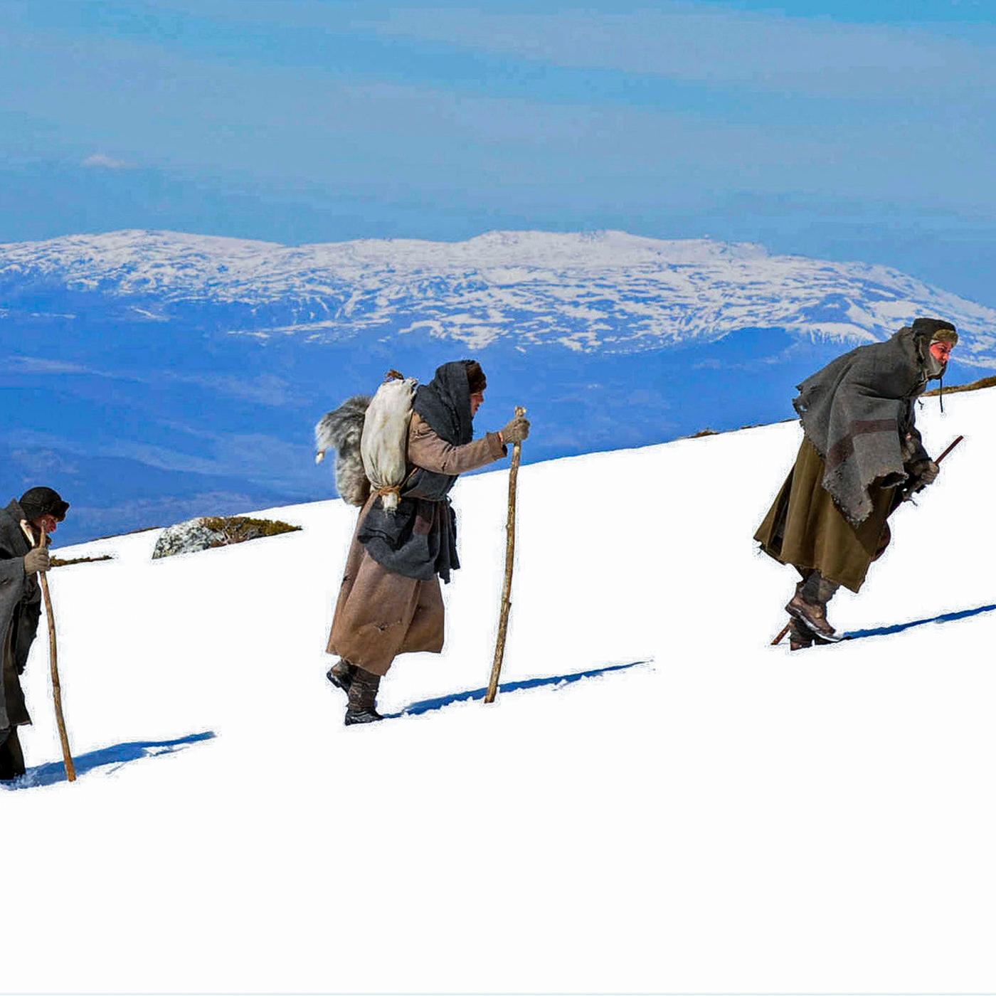 Vier Menschen, mit Decken geschützt und mit Stöcken in den Händen, wandern hintereinander einen schneebedeckten Berg hoch
