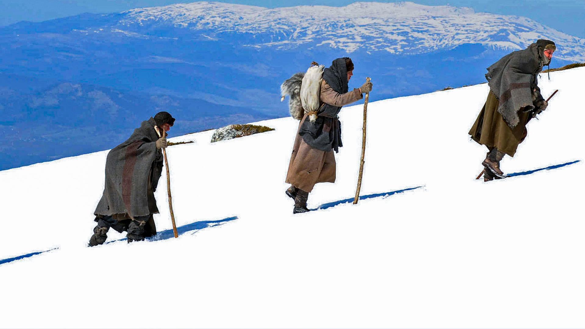 Vier Menschen, mit Decken geschützt und mit Stöcken in den Händen, wandern hintereinander einen schneebedeckten Berg hoch