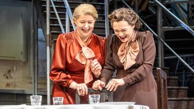 Bühnenszene: Zwei Frauen stehen vor einem gedeckten Tisch und lachen.