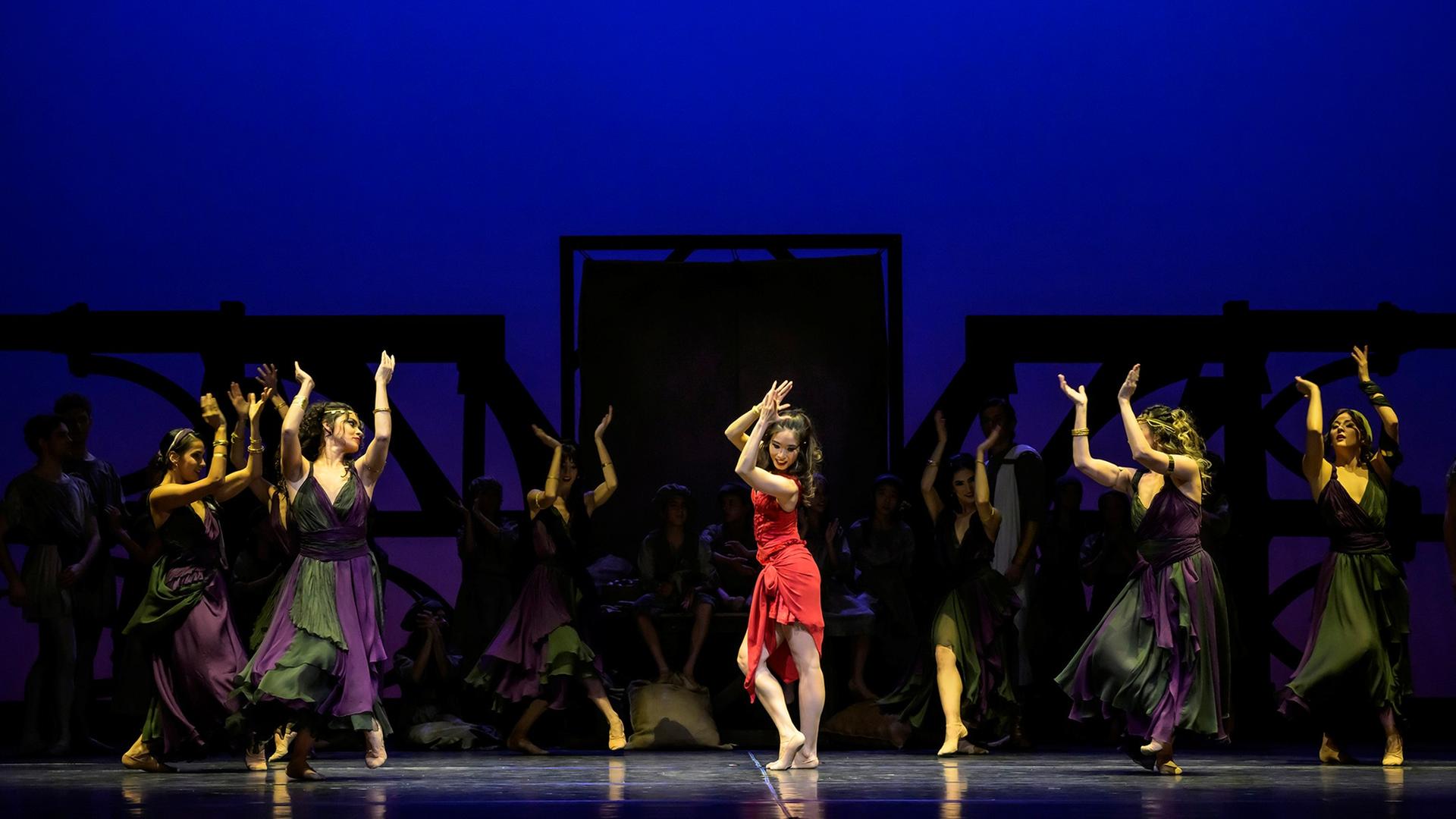 Tänzerinnen in bunten Kostümen tanzen auf der Bühne vor blauem Hintergrund.