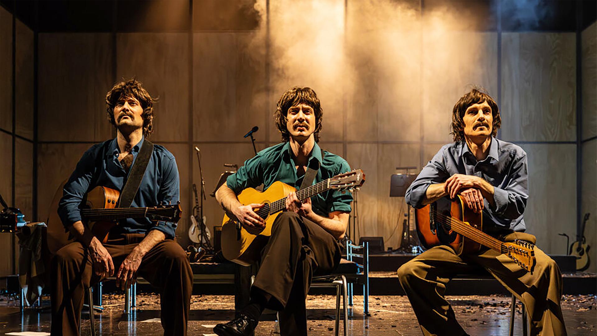 Bühnenszene: Drei Männer mit Gitarren, die Bekleidung der 1970er Jahre tragen, sitzen auf Stühlen.