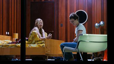 Bühnenszene: Eine Frau sitzt auf einem Bett und spricht mit einer anderen Frau, die auf einem Sessel davor sitzt.