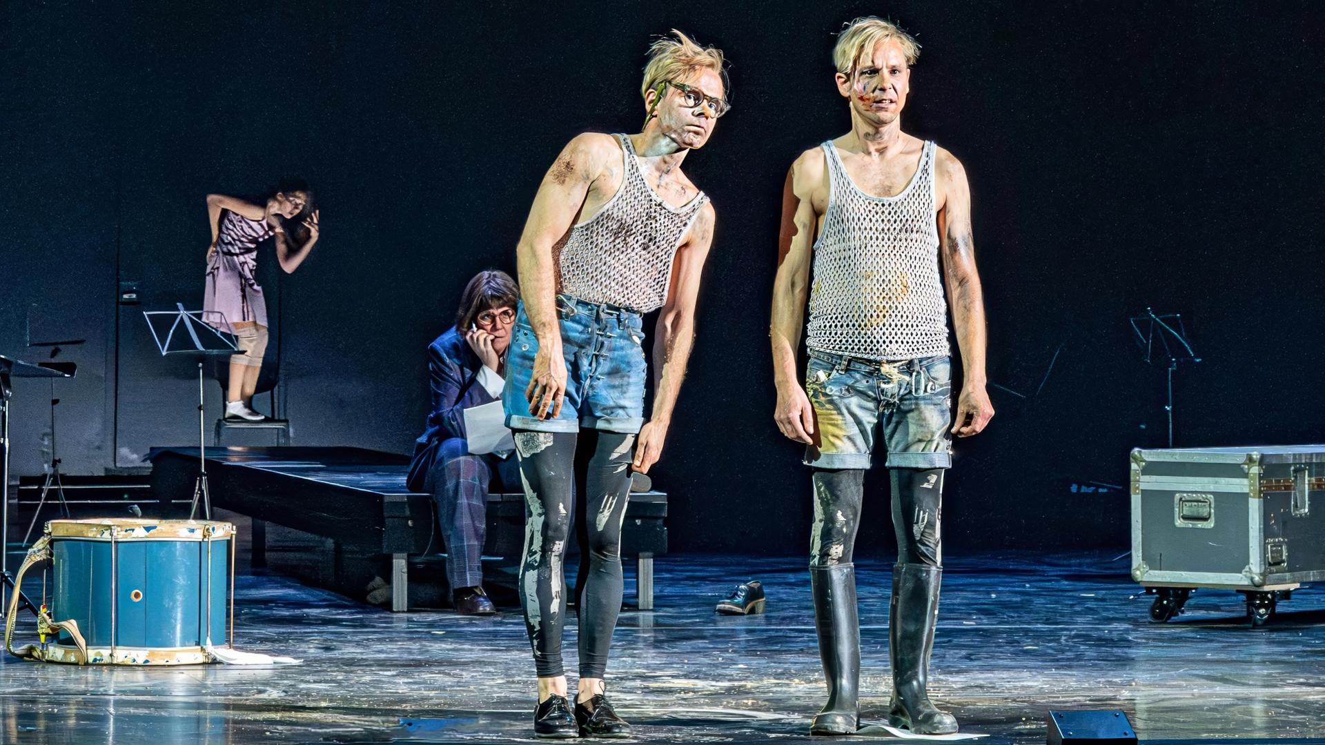 Zwei Männer in kurzen Hosen stehen auf einer Bühne. Im Hintergrund befinden sich zwei weitere Personen und Musikinstrumente.