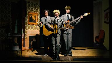 Drei Personen stehen in den gleichen grauen Anzügen mit schwarzer Krawatte auf einer Bühne. Die linke Person hält ein Tamborin in der Hand und die Person in der Mitte steht vor einem Mikrofon und singt hinein. Dabei spielt sie Gitarre. Die Person, die rechts steht, spielt einen E-Bass.