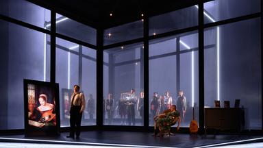 Bühnenszene: Man sieht die große Glasfensterfront eines Raumes im Halbdunkel. Links im Raum steht ein Mann neben einem Renaissancegemälde, rechts im Bild sitzt eine Frau in einem Sessel, neben einer Kommode. Draußen vor den Fenstern stehen Menschen und schauen hinein.