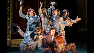 Bühnenszene: Drei Personen knien vor vier stehenden Personen. Alle tragen karierte Anzüge und halten Zylinderhüte in die Luft.