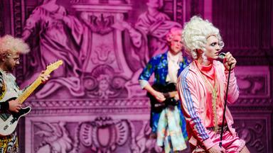 Bühnenszene: Eine Rockband, die Rokoko-Perücken und bunte Sportkleidung trägt spielt vor barocker Kulisse.