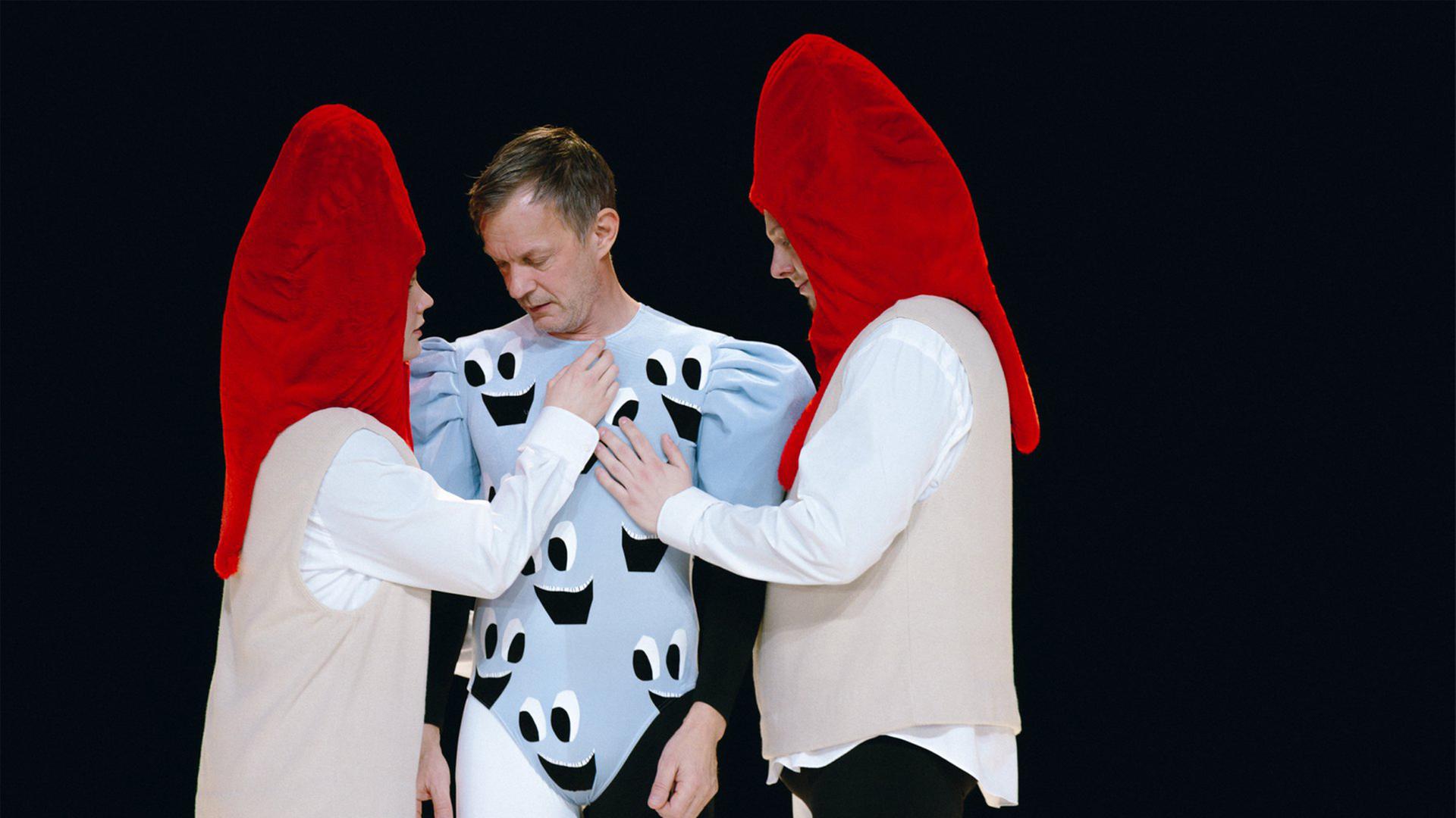 Bühnenszene: Ein Mann in hellblauem Turnanzug ist umgeben von zwei Personen, die rote Kopfbedeckungen tragen.