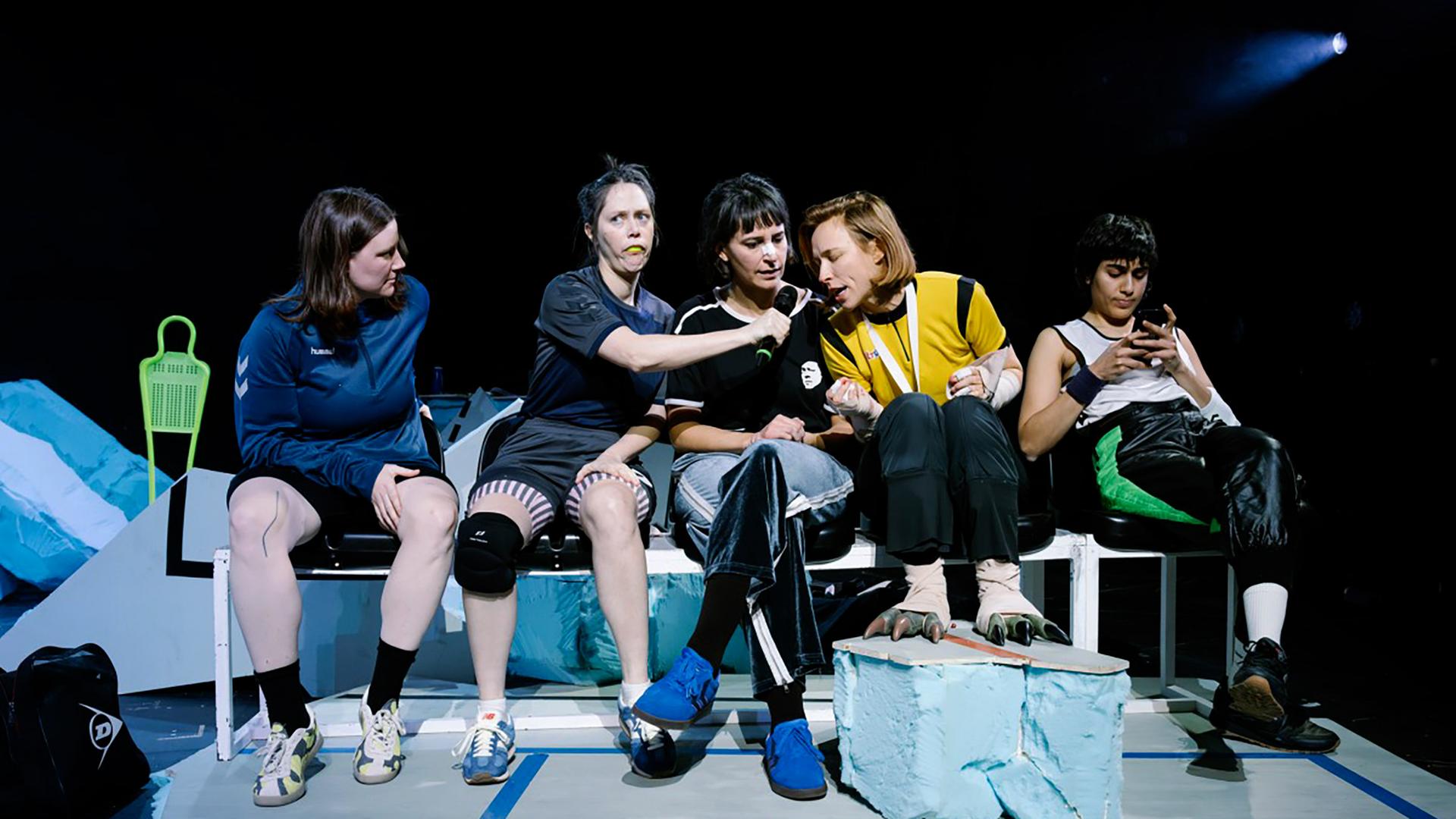 Bühnenszene: Frauen in Sportkleidung sitzen auf einer Bank.