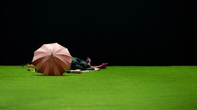 Bühnenszene: Eine Person liegt hinter einem geöffneten Regenschirm auf einem Kunstrasen.