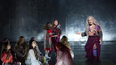 Bühnenszene: Mehrere Frauen in bunter Bekleidung sitzen und stehen auf der Bühne vor grauem Hintergrund.