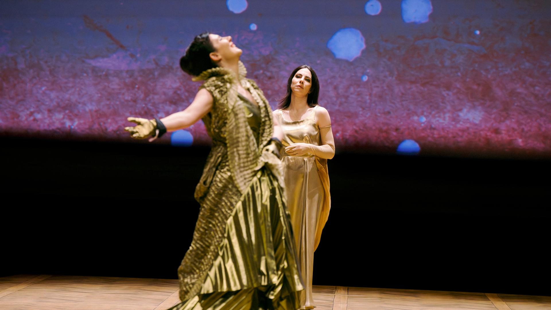 Bühnenszene: Zwei Frauen in goldenen Kleidern tanzen und stehen auf der Bühne.