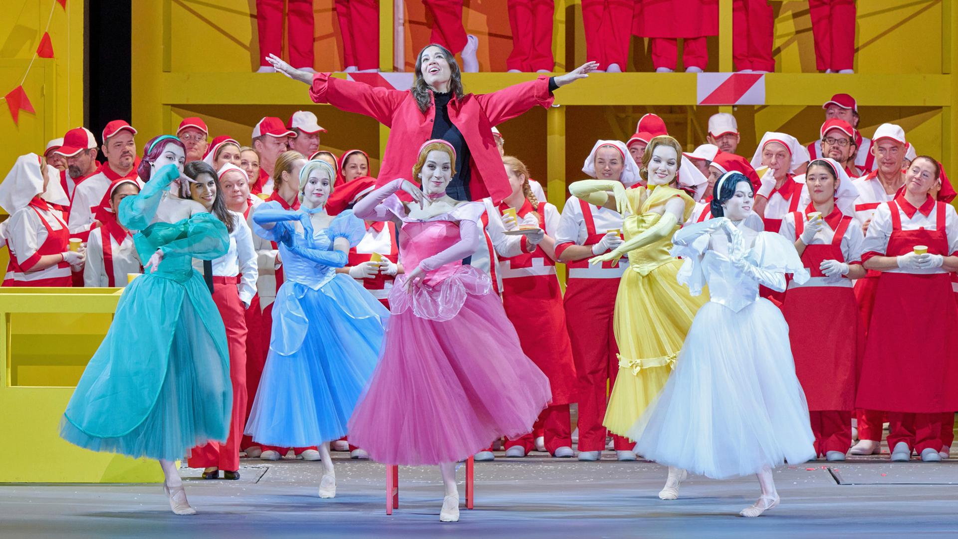 Bühnenszene: Frauen in bunten Kleidern tanzen. Im Hintergrund stehen Menschen in weiß-roter Bekleidung auf einer Empore.