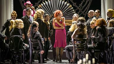 Bühnenszene: Eine Frau in einem pinkfarbenen Kleid ist umringt von Männern und Frauen.
