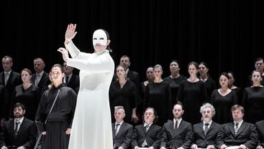 Bühnenszene: Man sieht eine weiß gekleidete Frau mit weißer Maske, die vor einer in schwarz gekleideten Menschenmenge steht.
